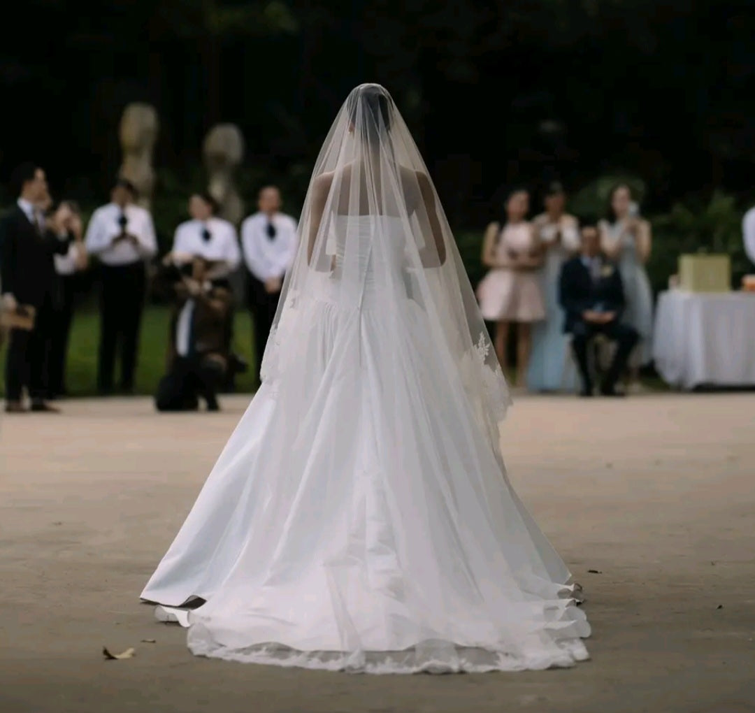 Bride in a white wedding dress with a long veil walking down the aisle outdoors with guests in the background.